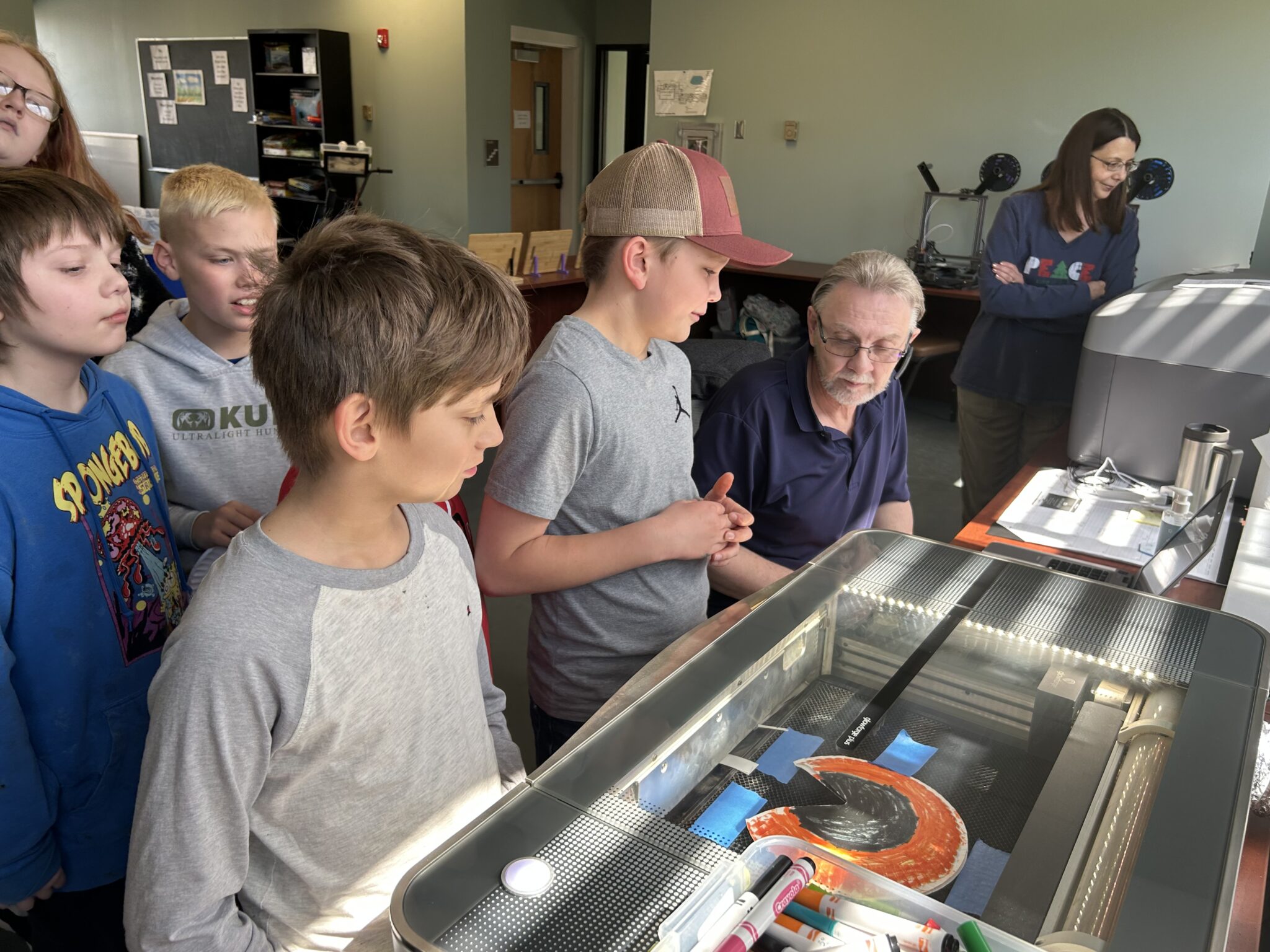 George Hebb, of The Computer Shop in Oakland, MD, works with students on the laser engraver to cut out eyeholes for eclipse glasses on student-designed paper plate sun masks.